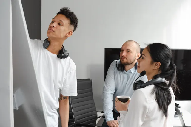 Confident multiethnic coworkers in white clothes with headsets in light workplace near chair and TV while looking at white board. Who owns what in a social listening team: six roles and their evidence